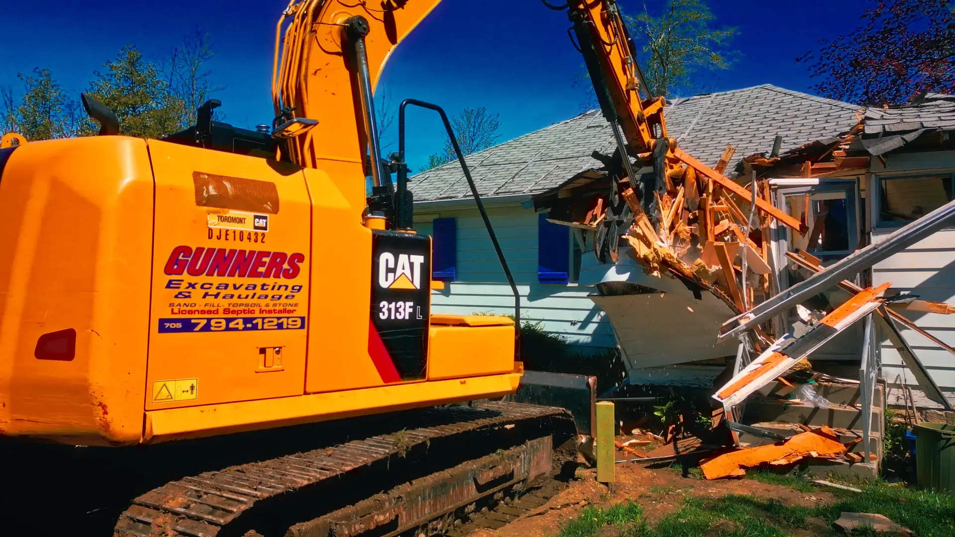 Branded excavator from a licensed septic installer demolishing a house with its bucket.